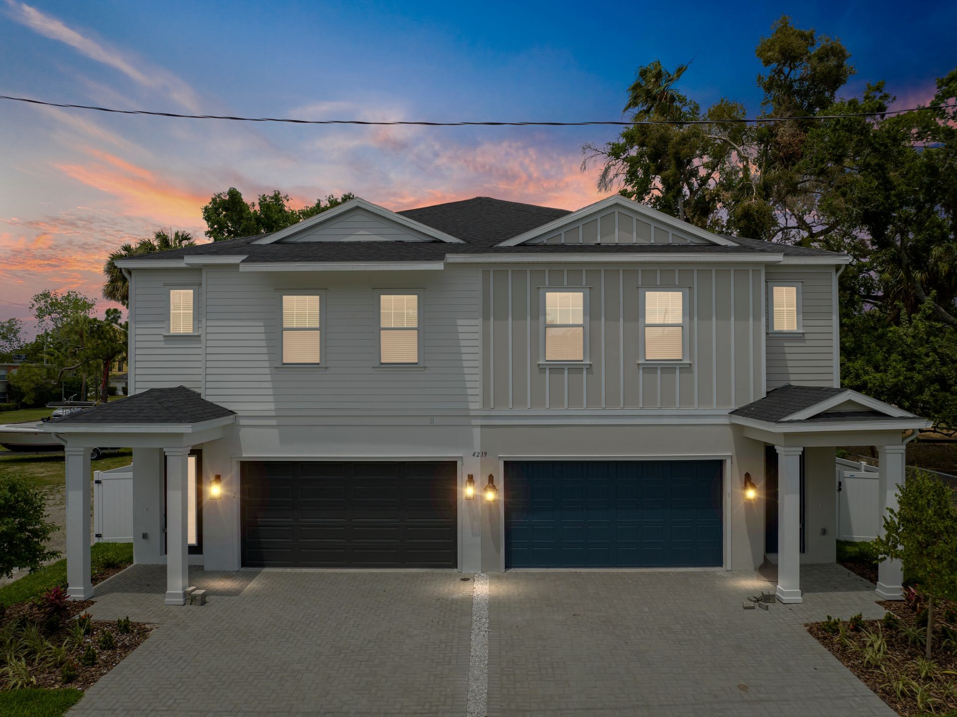 Front exterior of a modern duplex house with two garages and large windows, illuminated at dusk.