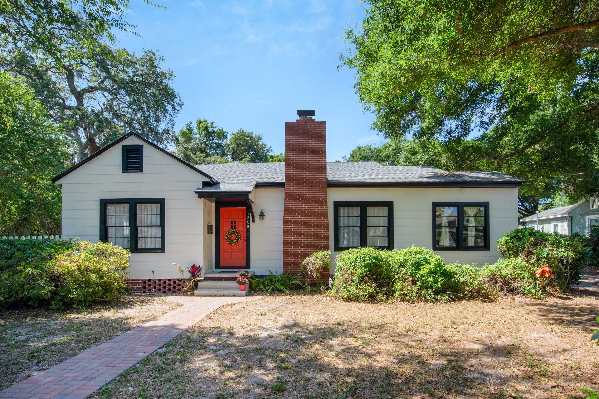 Single-story house with red door, brick chimney, surrounded by greenery under a clear blue sky.